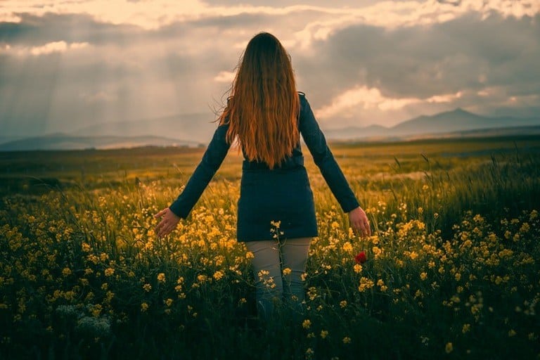 Woman in field of yellow flowers and sun rays streaming down from clouds in front of her